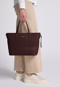 Persona sujetando un bolso tote Lazarotti de cuero marrón oscuro, vistiendo pantalones de pierna ancha beige y zapatillas de plataforma en color crema, de pie sobre un fondo claro.
