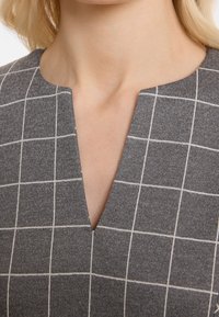 Grijze jurk met patroon met een v-hals, gemaakt van gestructureerde stof met een ruitpatroon in witte lijnen. Focus op de details van de halslijn.
