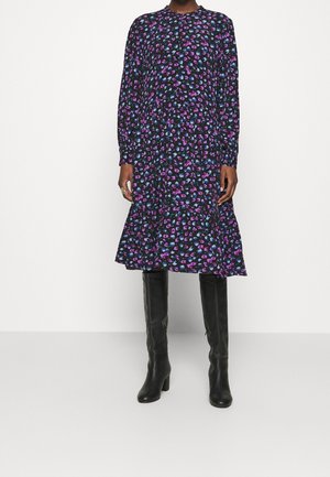 Femme portant une robe noire à manches longues avec un imprimé floral violet et bleu, associée à des bottes en cuir noires montant jusqu'aux genoux.