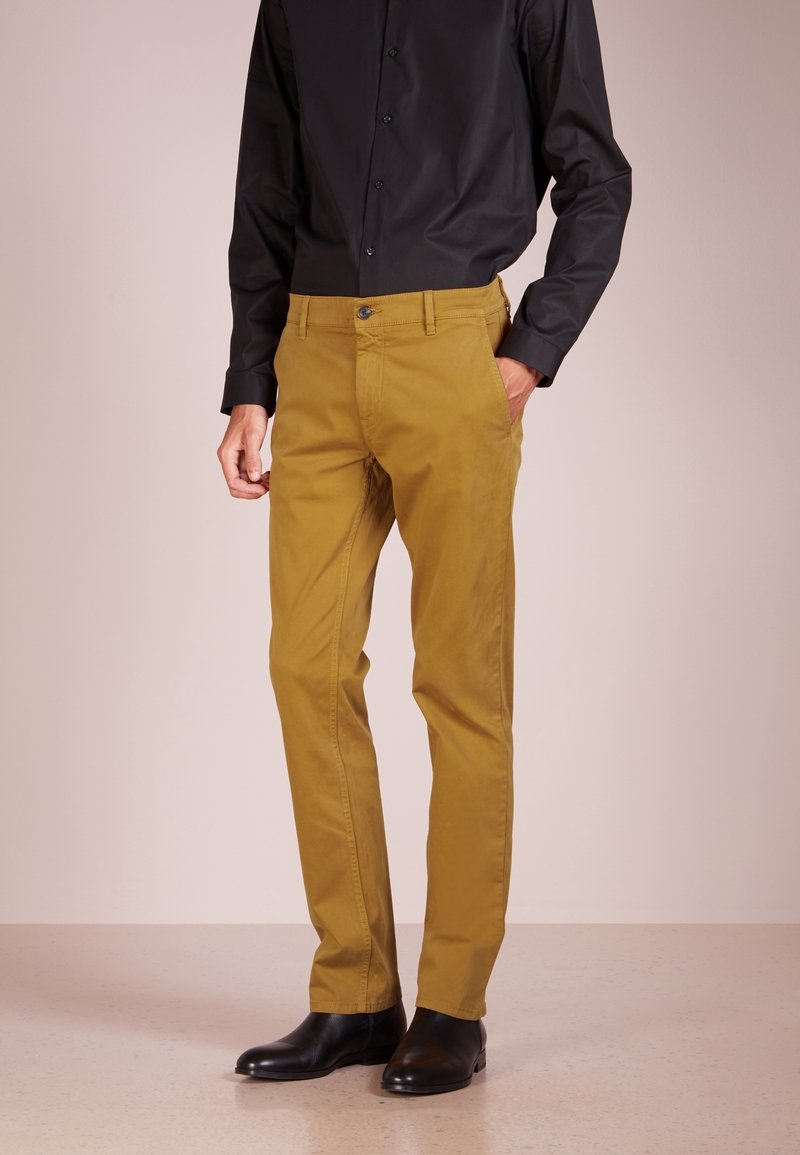 Jasnobrązowe spodnie o slim fit, z normalnym pasem, kieszeniami z przodu i zapięciem na guzik. Noszone z czarną bluzką z długim rękawem.