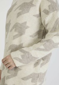 Manche de pull en maille beige et gris clair à motifs sur le bras d'une personne, avec une coupe décontractée.