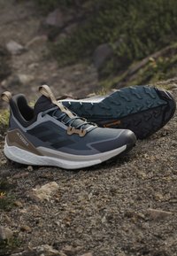 Een paar grijs-zwarte Adidas Terrex trailrunning-schoenen met robuuste Continental rubberzolen die op rotsachtige buitengrond rusten.