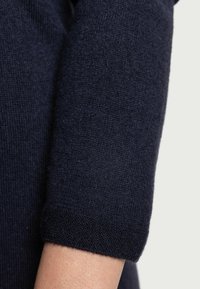 Primo piano di un avambraccio di una persona che indossa una manica di un maglione a maglia strutturata di un blu navy scuro che termina appena sotto il gomito.