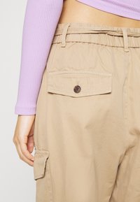 Calças cargo bege com cintura elástica, um bolso traseiro com botão e bolsos laterais, combinadas com um top canelado lavanda.