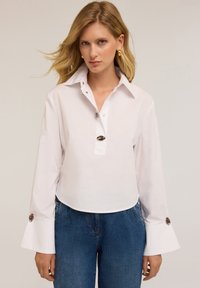 Camicia bianca di cotone con colletto a punta, maniche a campana lunghe, una patta con tre bottoni e dettagli decorativi con bottoni sui polsini delle maniche.