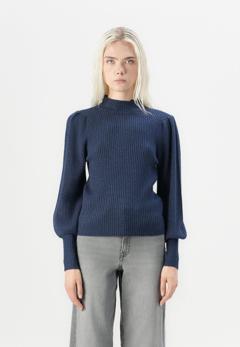 Navy ribbed knit sweater with a high neck and puffed long sleeves, paired with light grey wide-leg jeans. Textured fabric detail visible.