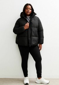 Mujer vestida con una chaqueta tipo puffer negra sobre una blusa gris y leggings negros, de pie de manera confiada frente a un fondo sencillo.