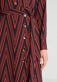 Abito geometrico con motivo a zigzag rosso e blu navy, maniche lunghe, chiusura con bottoni, cintura in vita e tessuto dalla texture liscia.