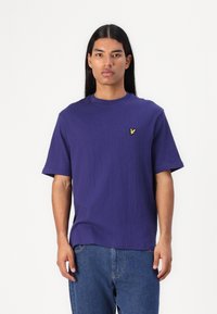 Camiseta corta de manga corta en color púrpura con un acabado texturizado, cuello redondo y un pequeño logo amarillo en el lado izquierdo del pecho. Combinada con jeans de mezclilla azules.