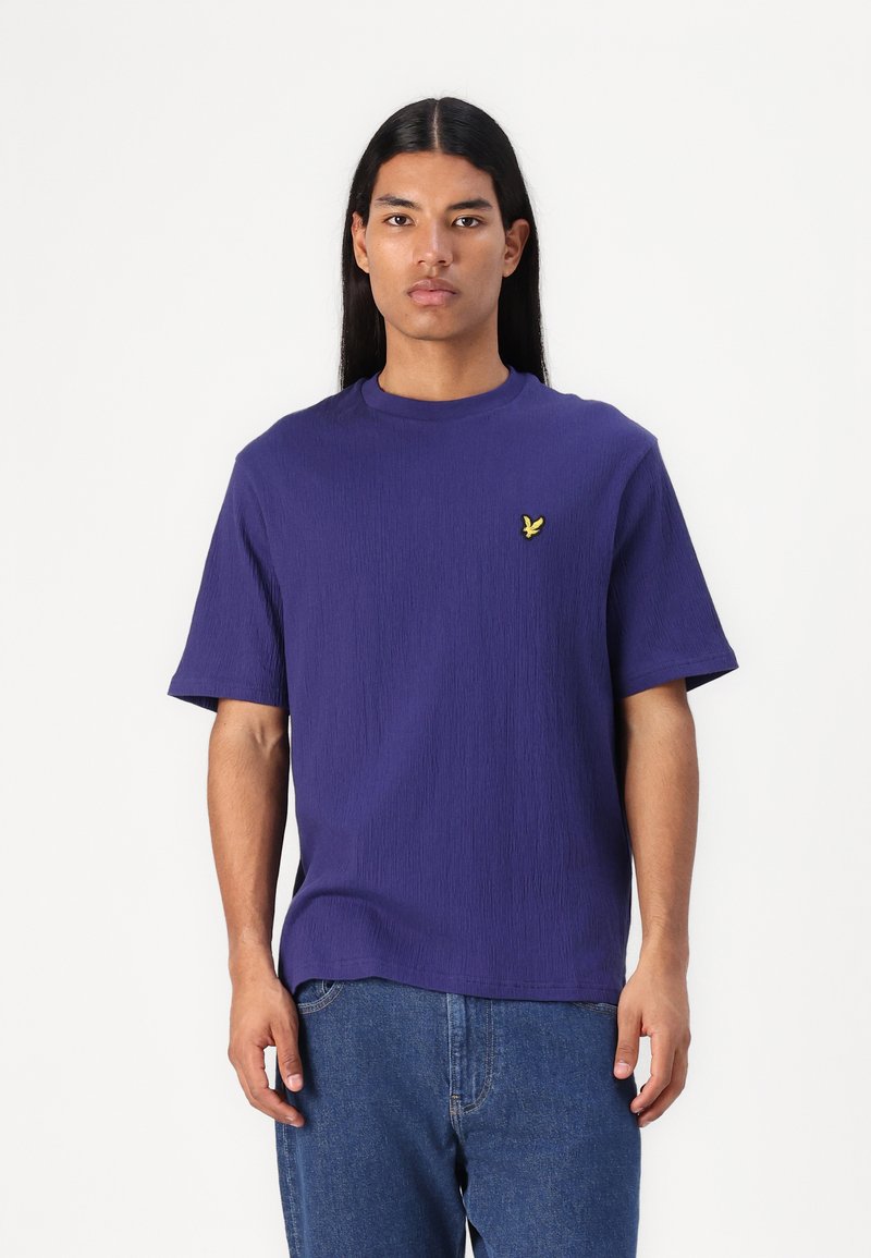 Camiseta corta de manga corta en color púrpura con un acabado texturizado, cuello redondo y un pequeño logo amarillo en el lado izquierdo del pecho. Combinada con jeans de mezclilla azules.
