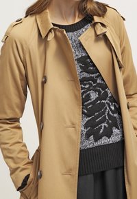 Beżowy płaszcz trench z zapięciem na guziki i naramiennikami, nałożony na czarno-biały sweter z wzorem liści. Gładka faktura.