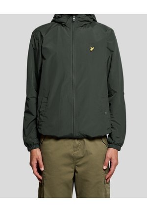Homme portant une veste à capuche vert foncé avec un petit logo d'oiseau jaune sur la poitrine et un pantalon cargo olive, debout les mains le long du corps.