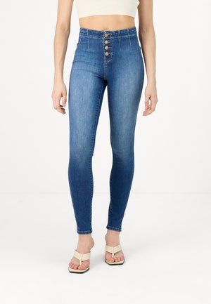 Kobieta ubrana w obcisłe niebieskie dżinsy z wysokim stanem z pięcioma widocznymi guzikami oraz beżowe sandały na obcasie z odkrytymi palcami, stojąca na białej podłodze.