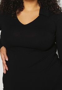 Top noir à manches longues avec un décolleté en V et un col, en tissu lisse, silhouette ajustée, avec des coutures ou des embellissements minimaux.