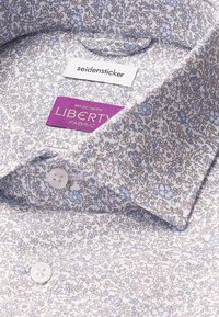 Camicia bianca con un delicato motivo floreale nei toni del blu e del rosa, dotata di colletto a punta e bottoni sulla pattina. Etichetta del tessuto inclusa.