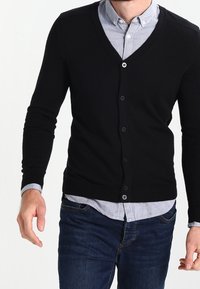 Svart v-neck cardigan gjord av ett mjukt material med långa ärmar, med knäppning framtill och en figursydd design, bärs över en ljusblå skjorta.