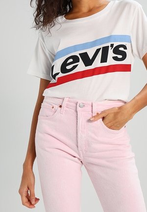 La camiseta blanca de Levi's con rayas rojas y azules presenta un gran texto en negro. Combinada con unos jeans rosa claro, que exhiben una tela suave y texturizada.