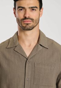 Chemise en lin marron à manches courtes avec un col en V, un devant boutonné et une poche poitrine. Le tissu a une texture apparente.
