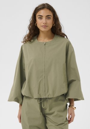 Femeie cu păr lung și creț, purtând o jachetă verde măsliniu, largă, cu fermoar și pantaloni asortați, pe un fundal simplu.