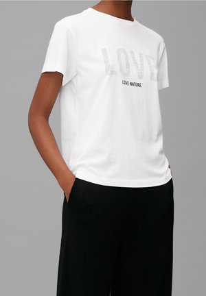 Personne portant un T-shirt blanc avec les inscriptions « LOVE » et « LOVE NATURE. », associé à un pantalon noir, mains dans les poches, devant un fond gris uni.