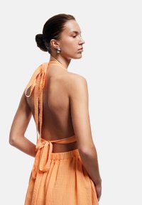 Robe orange à dos nu avec un design de halter-top, présentant un tissu texturé, de longues attaches au cou et à la taille, et un détail de jupe plissée.