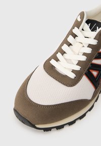 Prim-plan cu o sneaker cu suprapuneri din suede maro, plasă albă, șireturi albe și un logo "A" negru și portocaliu pe lateral.