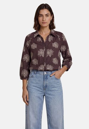 Femme aux cheveux foncés portant une chemise marron boutonnée avec des motifs floraux blancs et un jean taille haute bleu clair, une main dans la poche.