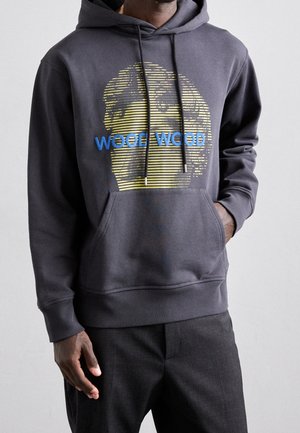 Man draagt een donkergrijze hoodie met een voorzak, blauwe tekst "WOOD WOOD" en een gele gestileerde gezichtsgrafiek gemaakt van horizontale lijnen.