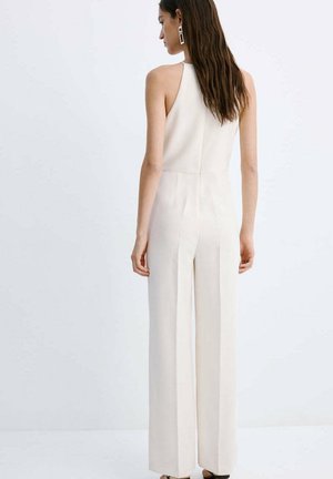 Mujer con cabello largo y castaño, vestida con un mono blanco sin mangas, de espaldas sobre un fondo blanco liso.