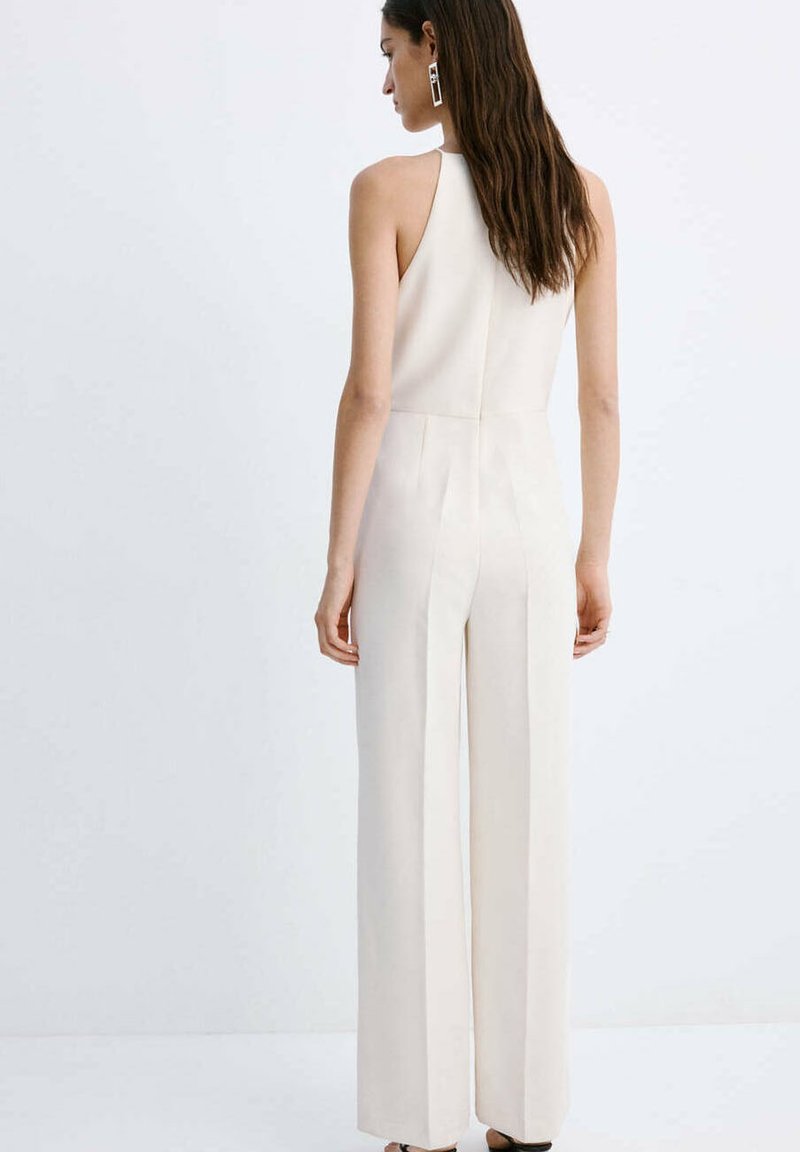 Femme aux longs cheveux bruns portant une combinaison blanche sans manches, de dos sur un fond blanc uni.