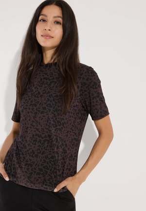 Frau mit langen dunklen Haaren, die ein braunes Kurzarmshirt mit schwarzem Leopardenmuster trägt, Hände in den Taschen, vor einem schlichten hellen Hintergrund stehend.