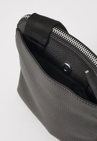 Sac en cuir noir texturé avec fermeture éclair et sangle en tissu. L'intérieur comporte une petite poche avec un bouton pression.