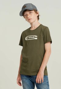 Oliven grøn bomulds t-shirt med korte ærmer og hvidt G-Star logo. Bæres med blå denim jeans og en grå kasket med et logo patch.