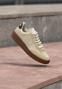 Zapatilla beige con suela marrón y lengüeta trasera negra, ligeramente elevada sobre una superficie de piedra rosada frente a un fondo de piedra gris.