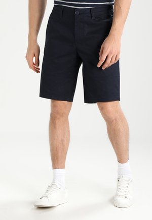Pantaloni corti in cotone blu navy con taglio dritto, chiusura a bottone e tasche laterali, abbinati a sneakers bianche e calzini bianchi visibili alla caviglia.