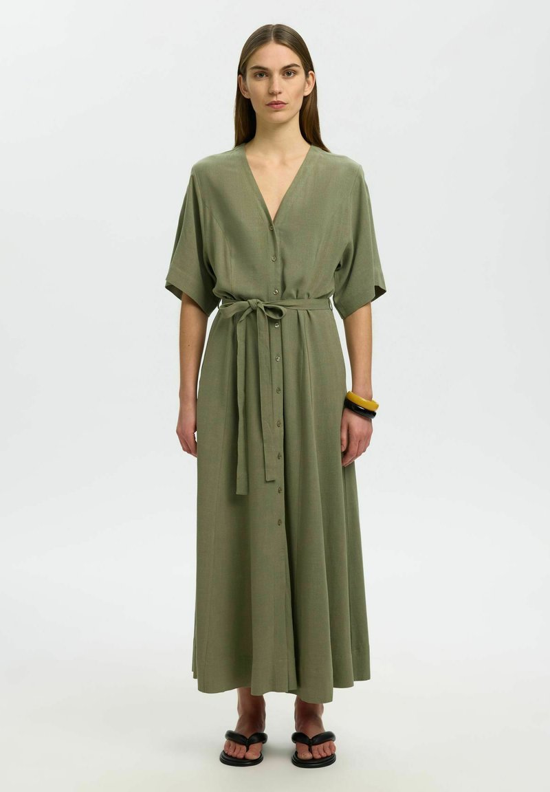 Femme debout portant une robe longue boutonnée vert olive avec une ceinture à la taille, et des bracelets noirs et jaunes à son poignet droit.