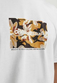 Hvid bomuld T-shirt med et rektangulært grafisk design af brune og gule blomster mønstre med teksten "Originals Studio" centreret på den.