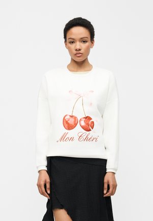 Γυναίκα που φοράει λευκό φούτερ με κόκκινο γραφικό κερασάκι και το κείμενο «Mon Chéri», σε συνδυασμό με μαύρη ασύμμετρη φούστα, στέκεται μπροστά σε λευκό φόντο.