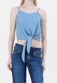 Top court en denim bleu clair avec fines bretelles, un détail noué à l'avant et une coupe décontractée. Associé à un jean en denim bleu foncé.