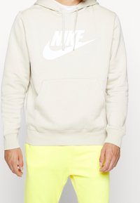 Světlé béžové mikinové tričko s kapsou na přední straně a bílým logem Nike, s kapucí na stažení a žebrovanými manžetami. Kombinováno s neonově žlutými kalhotami.