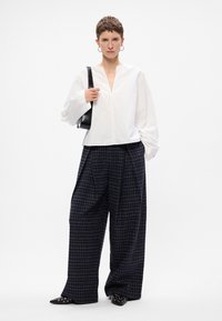 Bijela bluzica dugih rukava s V-izrezom, uparena s tamno kariranim širokim hlačama. Dodaci uključuju crnu torbu preko ramena i crne cipele s oštrim vrhom.