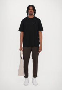 Tricou negru cu mânecă scurtă, croială relaxată, din material de bumbac. Purtați-l cu pantaloni maro și o geantă tote ușor bej; stând pe un fundal neutru.