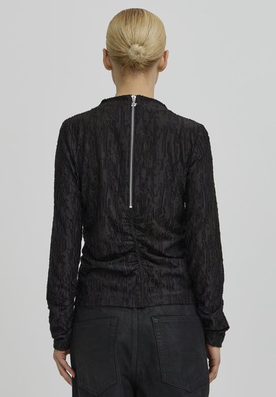 Fekete felső texturált anyaggal, hosszú ujjakkal és húzott dizájnnal a hátoldalon. Ezüst cipzár záródással a nyaknál.