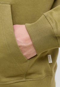 Oliwkowy sweter z kieszenią kangurka, żeberkowymi mankietami i szwami bocznymi. Widoczna miękka tekstura, z małą etykietą marki po boku.