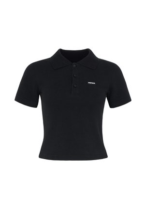 Femme debout portant un polo noir ajusté, un pantalon de survêtement gris ample avec des poignets élastiques, et des mocassins noirs sans lacets, sur un fond gris clair.