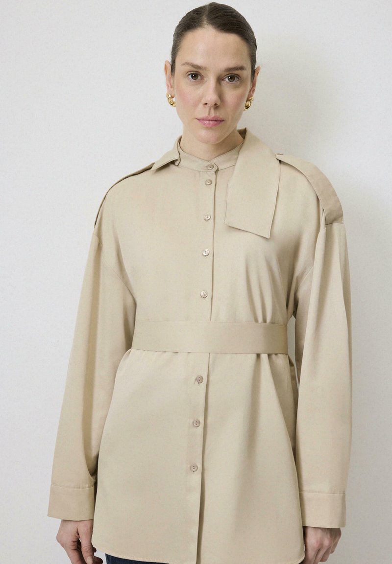 Camisa de manga larga beige con botones delanteros, cintura ceñida y cuello ancho. Tejido suave con una textura lisa y detalles de diseño minimalistas.