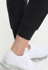 Zwarte sportbroeken met geperforeerde textuur op de dijen, gecombineerd met witte mesh sneakers met rubberen zolen. Close-up van het enkelgebied.