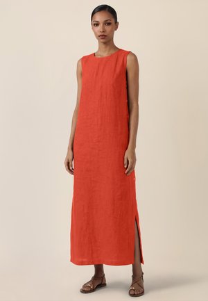 Frau trägt ärmelloses, knöchellanges, leuchtend orangefarbenes Kleid mit seitlichem Schlitz, steht mit neutralem Gesichtsausdruck und Sandalen vor beigem Hintergrund.