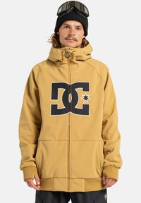 Man in een mosterdgele hoodie met een groot zwart-wit DC-logo, zwarte beanie en skibril op het hoofd, tegen een effen achtergrond.