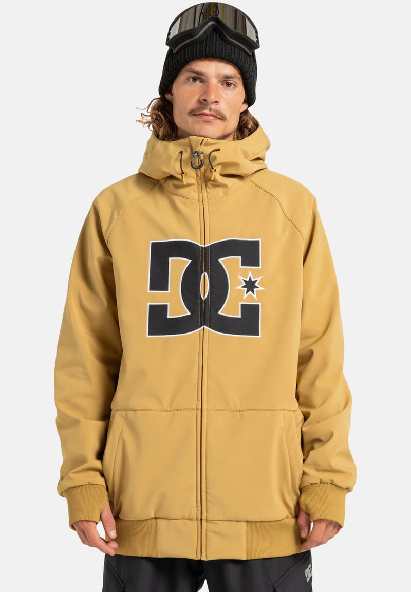 Man in een mosterdgele hoodie met een groot zwart-wit DC-logo, zwarte beanie en skibril op het hoofd, tegen een effen achtergrond.